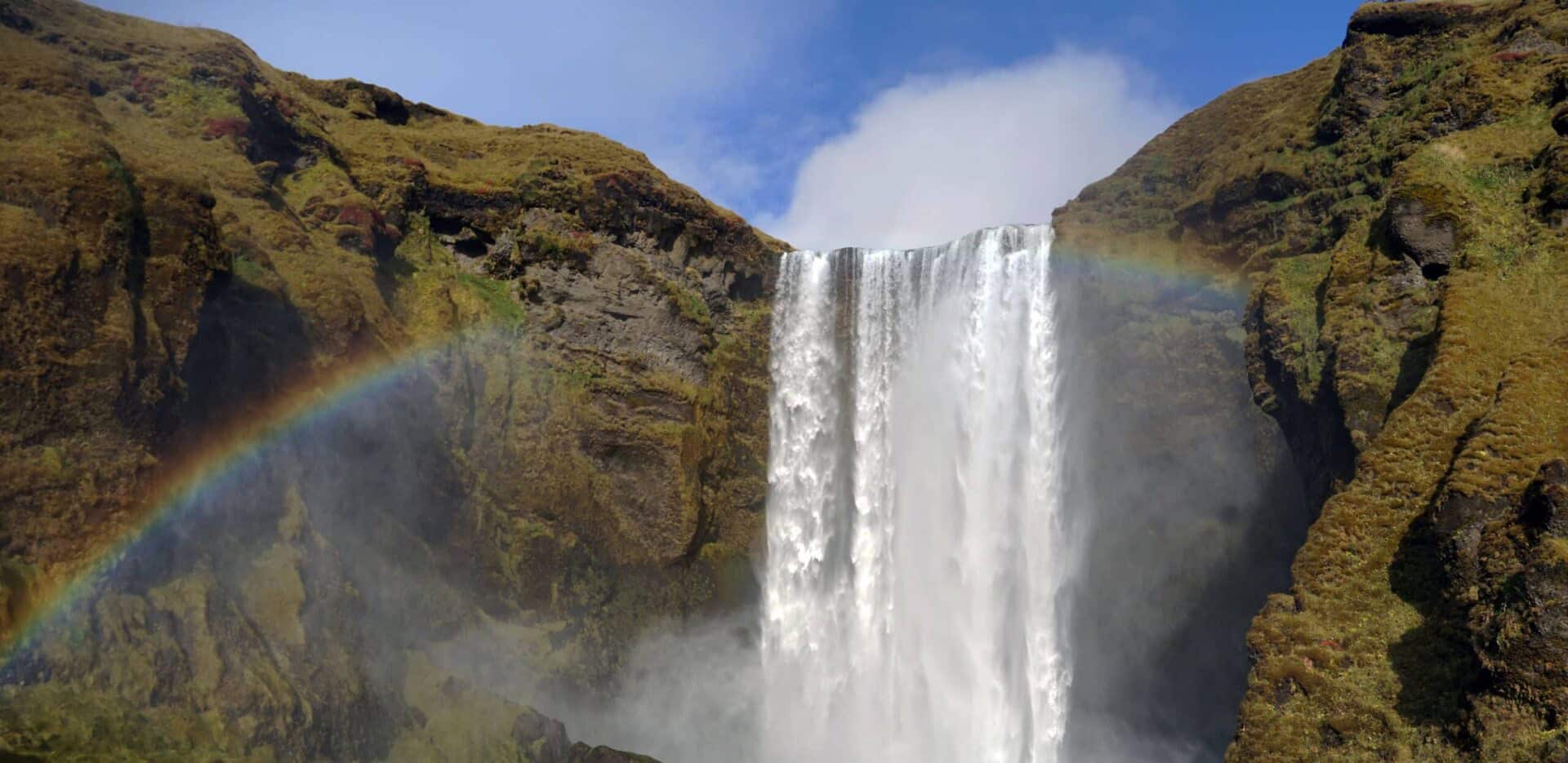 Skógafoss waterfall in southern Iceland cascading 200 feet between green volcanic cliffs under a blue sky, with a vivid rainbow stretching across the misty pool below. Made famous in Game of Thrones.