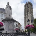 Bronze statue of Margaret of Austria framed by hanging pink petunias with St. Rumbold's Tower behind