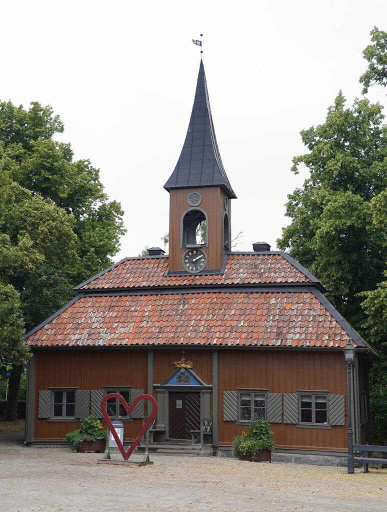 Oversized red heart sculpture in front of Sigtuna's tiny 1744 Town Hall, Swedish flags flying, gravel square in the foreground.