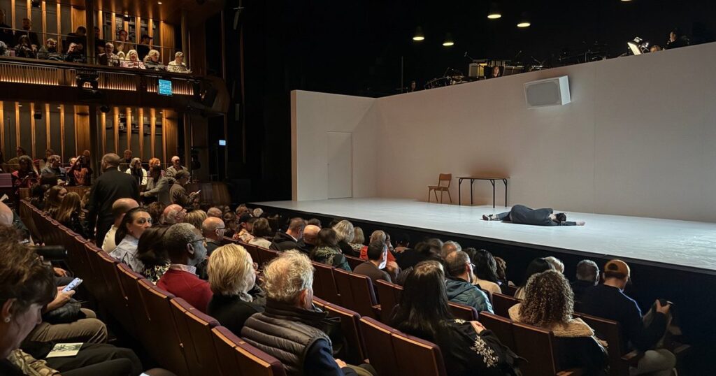 4.48 Psychosis review photo of a performer lying on a stark white stage as the Linbury Theatre audience fills the seats.