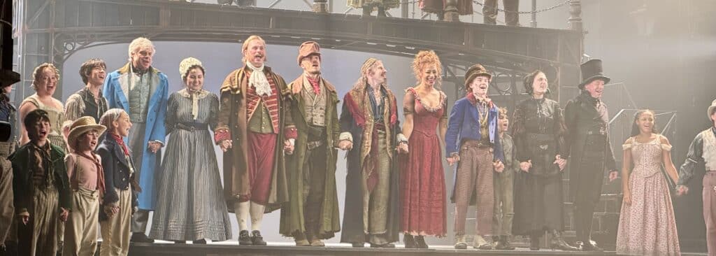 liver West End review photo of the cast bowing beneath an iron footbridge lined with Victorian lamp posts on the Gielgud Theatre stage.