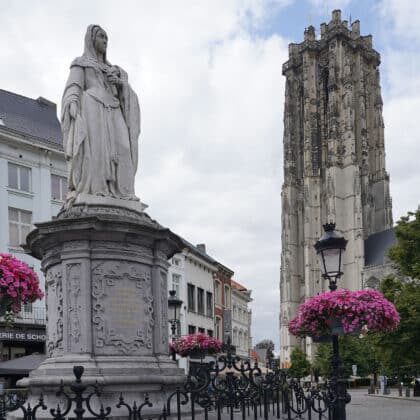Bronze statue of Margaret of Austria framed by hanging pink petunias with St. Rumbold's Tower behind